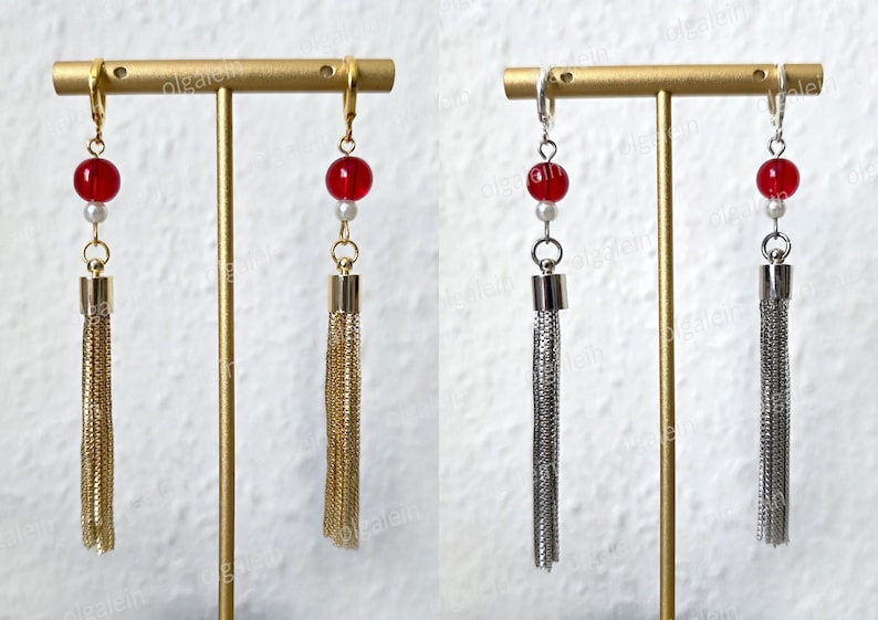 May include: Earrings featuring a red bead, a small white bead, and a chain tassel. One pair has gold-coloured metal, and the other has silver-coloured metal. The earrings are displayed on a gold-coloured stand.