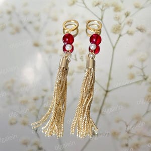 May include: A pair of gold-coloured tassel earrings. Each earring has a gold hoop, red beads, and a gold tassel. The earrings are shown against a soft, blurred background with white flowers.