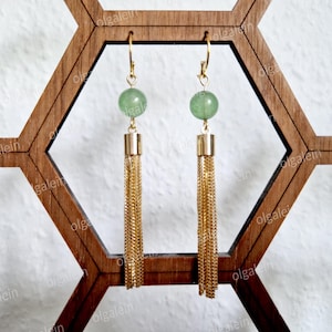May include: A pair of gold-toned earrings with green gemstone beads and long chain tassels. The earrings are displayed on a wooden geometric stand, showcasing their elegant design and craftsmanship. The earrings are a stylish accessory.