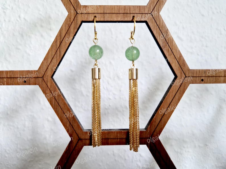 May include: A pair of gold-toned earrings with green gemstone beads and long chain tassels. The earrings are displayed on a wooden geometric stand, showcasing their elegant design and craftsmanship. The earrings are a stylish accessory.
