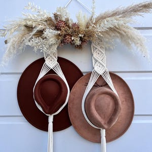 May include: A macrame hat holder with two hats. The hats are brown and tan, suspended by white macrame. The holder is decorated with dried flowers and pampas grass. The background is a white wooden wall.