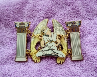 May include: A gold-colored enamel pin featuring a blindfolded figure with angel wings, chained between two white columns with blue and gold accents. The figure is dressed in white and seated. The pin is on a purple background.