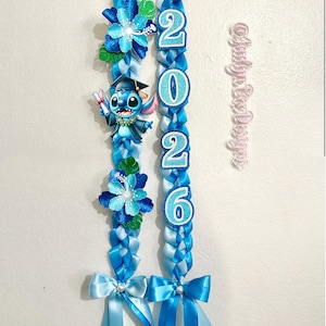 May include: Two blue graduation leis with a Stitch character, flowers, and the year 2026. The leis are made of braided ribbons and adorned with bows. A pink, cursive text is also visible.