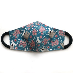 May include: A teal blue face mask with a repeating pattern of pink flowers and white koalas. The mask has black elastic ear loops.