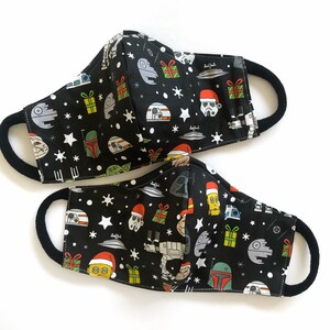 Op de afbeelding: Twee zwarte stoffen mondmaskers met een kerst- en Star Wars-thema. De maskers bevatten verschillende personages en symbolen, waaronder de kerstman, Darth Vader en de Millennium Falcon. De maskers zijn beide omkeerbaar, met een ander ontwerp aan elke kant.