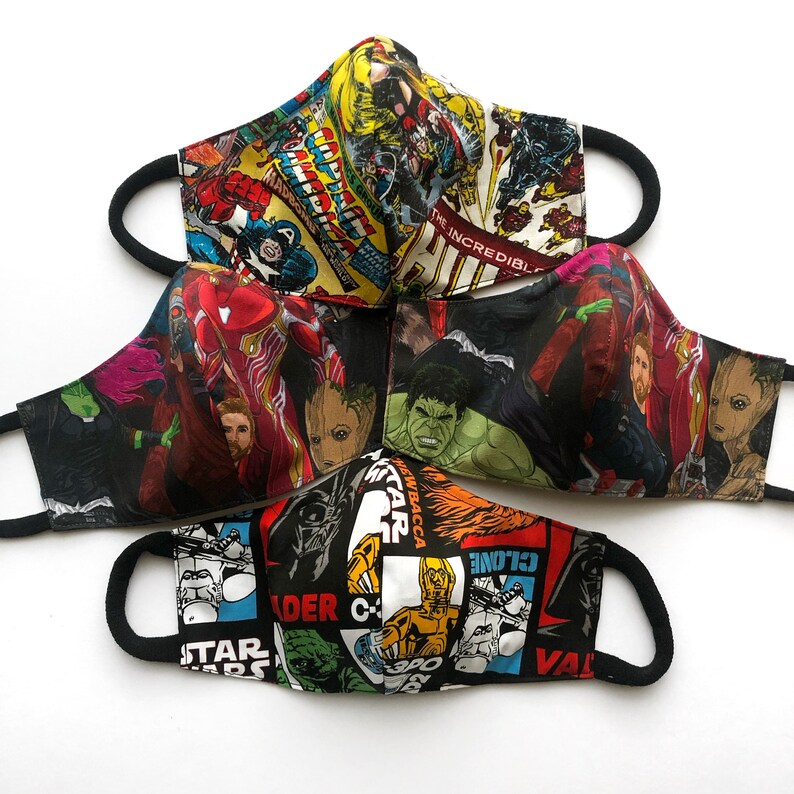Op de afbeelding: Vier verschillende stoffen mondmaskers met verschillende superhelden- en stripfigurenontwerpen. De maskers zijn zwart, rood, blauw en groen met afbeeldingen van personages zoals Iron Man, Hulk en Darth Vader.