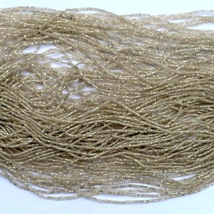 May include: A large quantity of small, clear glass beads with a subtle iridescent sheen. The beads are arranged in a pile and a smaller strand of the beads is shown next to a US dime for scale.