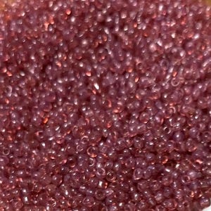 May include: A clear glass bowl filled with small, translucent, reddish-purple seed beads. The beads are densely packed, creating a textured surface. The bowl is set against a light brown background, highlighting the beads' color and shine. Ideal for jewelry making.