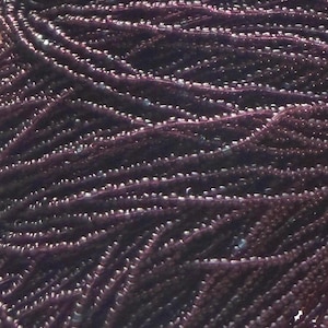 May include: Close-up of a collection of dark purple seed beads. The beads are strung together, creating a textured, flowing appearance. The beads are small and round, with a slightly iridescent sheen.