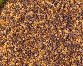 May include: A large collection of small, round, translucent glass seed beads in varying shades of amber, honey, and dark brown. Each bead features a central hole, creating a uniform, textured surface of mixed warm-toned, earthy colors.