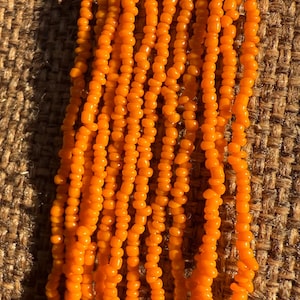 May include: Close-up of strands of orange beads. The beads are small, round, and strung together, creating a textured, flowing appearance. The background is a rough, woven fabric, providing a contrasting backdrop to the vibrant beads. The beads are a warm, bright orange.
