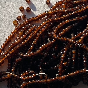 May include: A large pile of brown seed beads. The beads are small and round, and they are arranged in a random pattern. The beads are shiny and have a slight iridescent sheen.