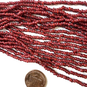 May include: A collection of small, round, deep red glass beads arranged in multiple parallel strands. The beads have a slightly translucent, glossy finish. A copper-colored United States one-cent coin is placed below the beads for scale.