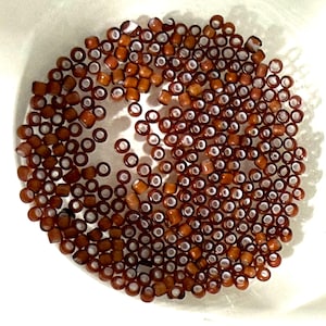 May include: A collection of small, brown, cylindrical beads with white centers. The beads are scattered in a circular pattern, creating a textured appearance. The beads are likely used for jewelry making or other crafts.