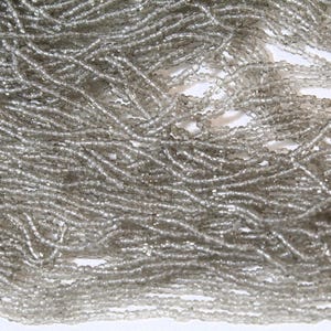 May include: Close-up of strands of small, translucent, gray beads. The beads are strung together, creating a flowing, textured appearance. The beads are likely used for jewelry making or other crafts.