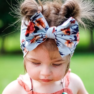 May include: A young girl with brown hair wearing a blue headband with a floral pattern and a coral colored dress with a multi-colored beaded necklace.