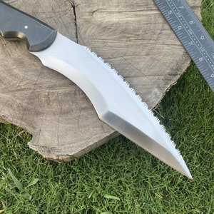 May include: A silver knife with a black handle and a serrated edge. The knife is resting on a wooden surface with green grass in the background.