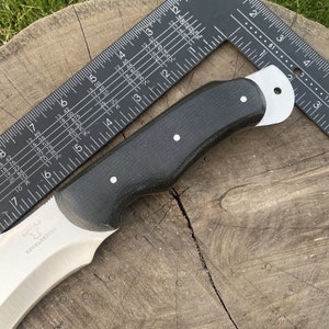 May include: A black handled knife with a curved blade and a silver finish. The blade has the text "ROVALKNIVES" etched into it. The knife is resting on a wooden surface with a ruler beside it.