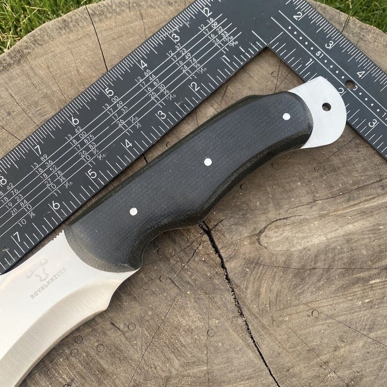 May include: A black handled knife with a curved blade and a silver finish. The blade has the text "ROVALKNIVES" etched into it. The knife is resting on a wooden surface with a ruler beside it.