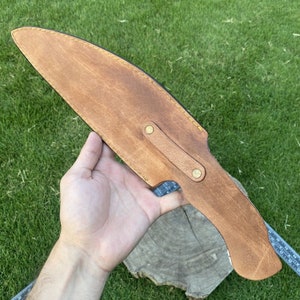 May include: A brown leather sheath with a flap and two brass snaps. The sheath is designed to hold a knife or other sharp object.