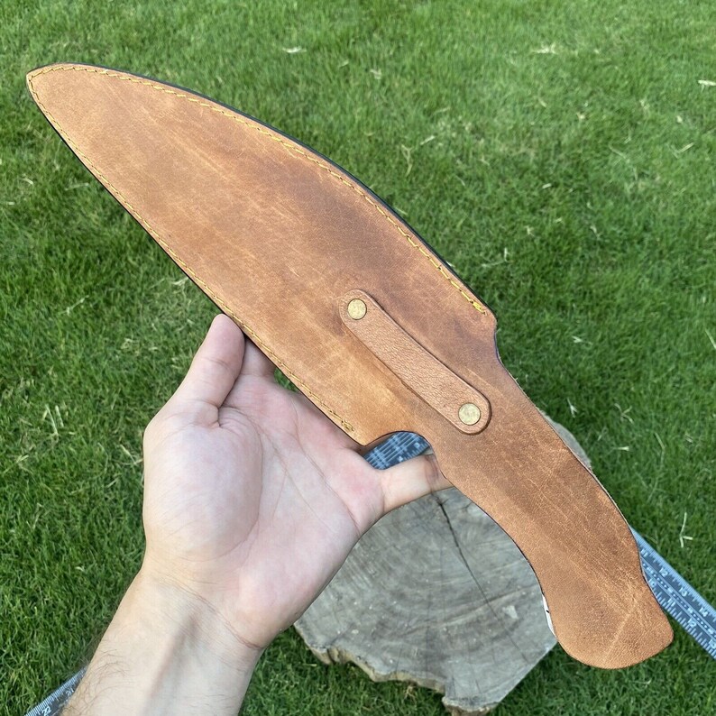 May include: A brown leather sheath with a flap and two brass snaps. The sheath is designed to hold a knife or other sharp object.