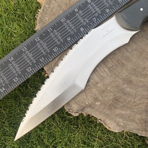 May include: A silver-colored knife with a serrated edge and a black handle. The knife is measured against a ruler, showing it is approximately 17 inches long. The knife has the text "ROVALKNIVES" printed on the handle.