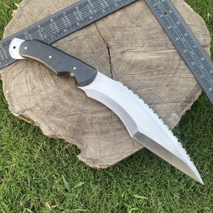 May include: A black and silver curved knife with a serrated edge and a black handle. The knife is resting on a wooden surface.