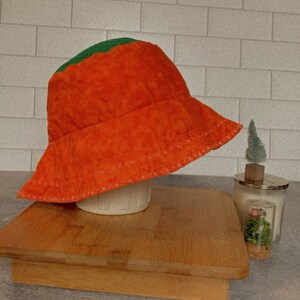 May include: Orange and green bucket hat with a stitched brim. The hat is sitting on a wooden cutting board.