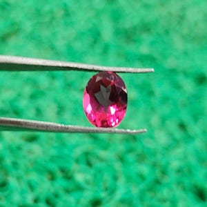 May include: A vibrant, oval-cut gemstone with a deep magenta-pink hue and multifaceted surface, held securely between the two thin, metallic prongs of a pair of tweezers. The stone displays clear internal reflections and a polished finish.