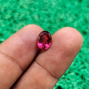 May include: A faceted oval-shaped gemstone with a vibrant pinkish-red hue. The stone features a clear, transparent quality with multiple geometric facets that reflect light, showcasing a brilliant, polished surface and a symmetrical cut.