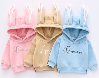 May include: Three hooded sweatshirts with bunny ears. The sweatshirts are pink, beige, and light blue. The pink sweatshirt has the name "Frances" written on it. The beige sweatshirt has the name "Ary" written on it. The light blue sweatshirt has the name "Roman" written on it.