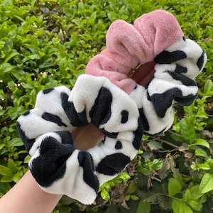 May include: Two black and white cow print scrunchies and one pink scrunchie. The scrunchies are made of soft, fuzzy fabric.