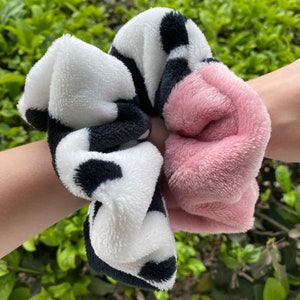 May include: Two fuzzy hair scrunchies. One is black and white with a cow print pattern. The other is a solid pink.