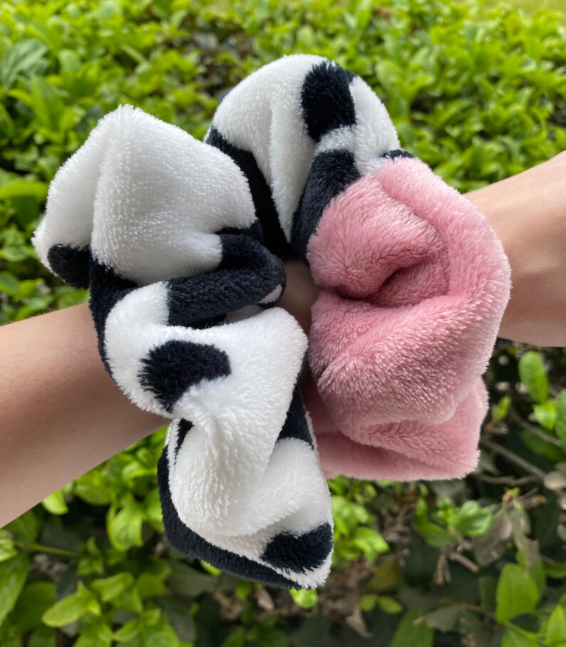 May include: Two fuzzy hair scrunchies. One is black and white with a cow print pattern. The other is a solid pink.