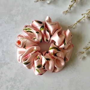 May include: A pale pink satin scrunchie featuring a floral design with green leaves and small orange flowers. The scrunchie is gathered, offering a soft, shiny look, ideal for hair accessories.