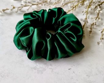 May include: A dark emerald green satin scrunchie. The hair accessory is made of a shiny, gathered fabric. The scrunchie is circular and has a soft, luxurious appearance. White flowers are in the background.