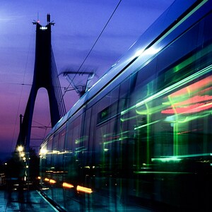 Könnte beinhalten: Ein moderner Tram fährt nachts über eine Brücke. Die Fenster des Trams spiegeln die Lichter der Stadt und die Brückenkabel wider. Der Himmel ist tiefblau mit einem Hauch von Lila. Das Bild trägt die Bildunterschrift "DAVID CLYNCH PHOTOGRAPHY".