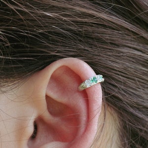 May include: Close-up of an ear adorned with a delicate ear cuff. The ear cuff is silver-toned and features a row of small, sparkling emerald and clear gemstones. The background includes brown hair.