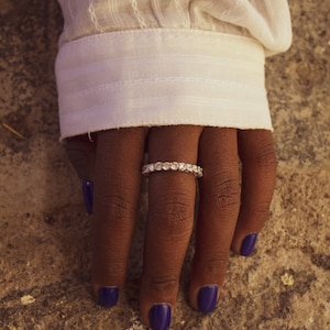 Può includere: Una mano dalla pelle scura con un anello d'argento con più pietre trasparenti. Le unghie sono dipinte di un viola intenso. Il polsino di una camicia bianca a maniche lunghe è visibile.