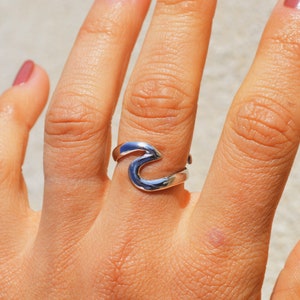 Può includere: Un anello d'argento a forma di onda su un dito con smalto rosa chiaro.