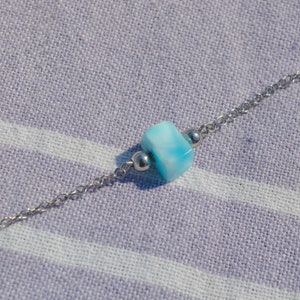 Könnte beinhalten: Eine zarte Halskette mit einer kleinen, hellblauen, würfelförmigen Perle mit weißer Marmorierung. Die Perle ist auf einer feinen Silberkette zentriert, mit einer kleinen Silberperle auf jeder Seite. Die Halskette liegt auf einem hellviolett-weiß gestreiften Stoff.