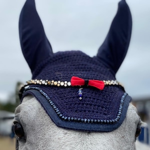 Puede incluir: Un gorro anti-moscas azul marino para caballo con un lazo rojo y una banda de frente adornada con cristales. El gorro se lleva en un caballo gris.
