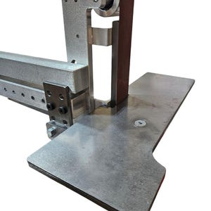 May include: A close-up of a metal belt sander with a brown sanding belt. The sander has a metal base and a metal arm that holds the sanding belt. The sanding belt is wrapped around a metal roller.