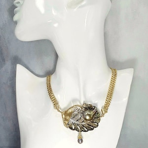 Peut inclure: Un collier en chaîne dorée avec un grand pendentif en forme de feuille avec une perle au centre. Le pendentif est en or, argent et métal noir.