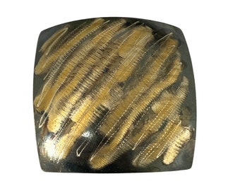 May include: Square, silver-toned metal object with a gold-coloured, textured design. The gold design is composed of diagonal, parallel lines that create a textured pattern across the surface. The object has rounded corners.