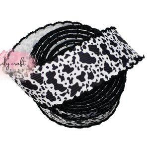 K&ouml;nnte beinhalten: Eine Rolle schwarz-wei&szlig;es Kuhfell-Printband mit gewelltem Rand. Das Band ist eng gewickelt und zeigt das sich wiederholende Muster schwarzer Flecken auf wei&szlig;em Grund. Das Logo von Lily Craft Supplies ist sichtbar.