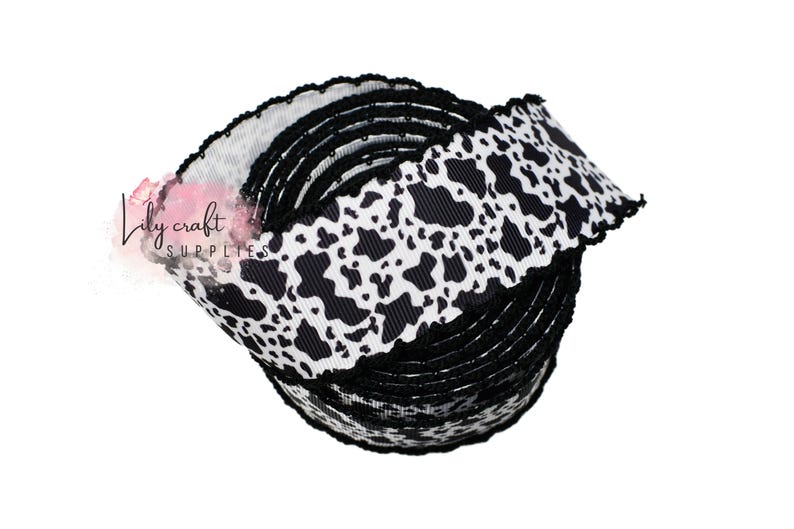 K&ouml;nnte beinhalten: Eine Rolle schwarz-wei&szlig;es Kuhfell-Printband mit gewelltem Rand. Das Band ist eng gewickelt und zeigt das sich wiederholende Muster schwarzer Flecken auf wei&szlig;em Grund. Das Logo von Lily Craft Supplies ist sichtbar.