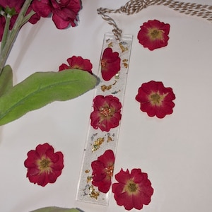 May include: A white bookmark with a clear resin panel featuring pressed red flowers and gold and silver glitter. The bookmark has a brown twine tassel.