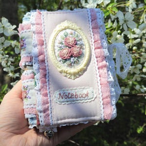 Könnte beinhalten: Ein rosa Stoffnotizbuch mit einem cremefarbenen ovalen Rahmen mit Blumendesign. Das Notizbuch ist mit rosa Rüschen und weißer Spitze verziert. Das Wort "Notebook" ist auf dem Einband gestickt.