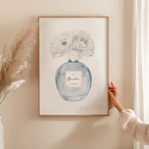 May include: Watercolor print of a perfume bottle with white flowers. The blue bottle has a label that reads "Belle" and "eau de parfum". The artwork is framed in a light brown frame.